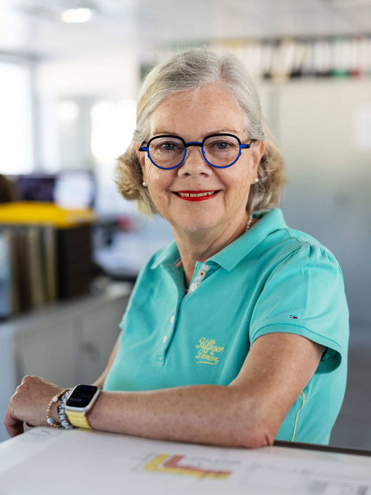 Frau im türkisfarbenen Shirt sitzt an einem Tisch und lächelt direkt in die Kamera