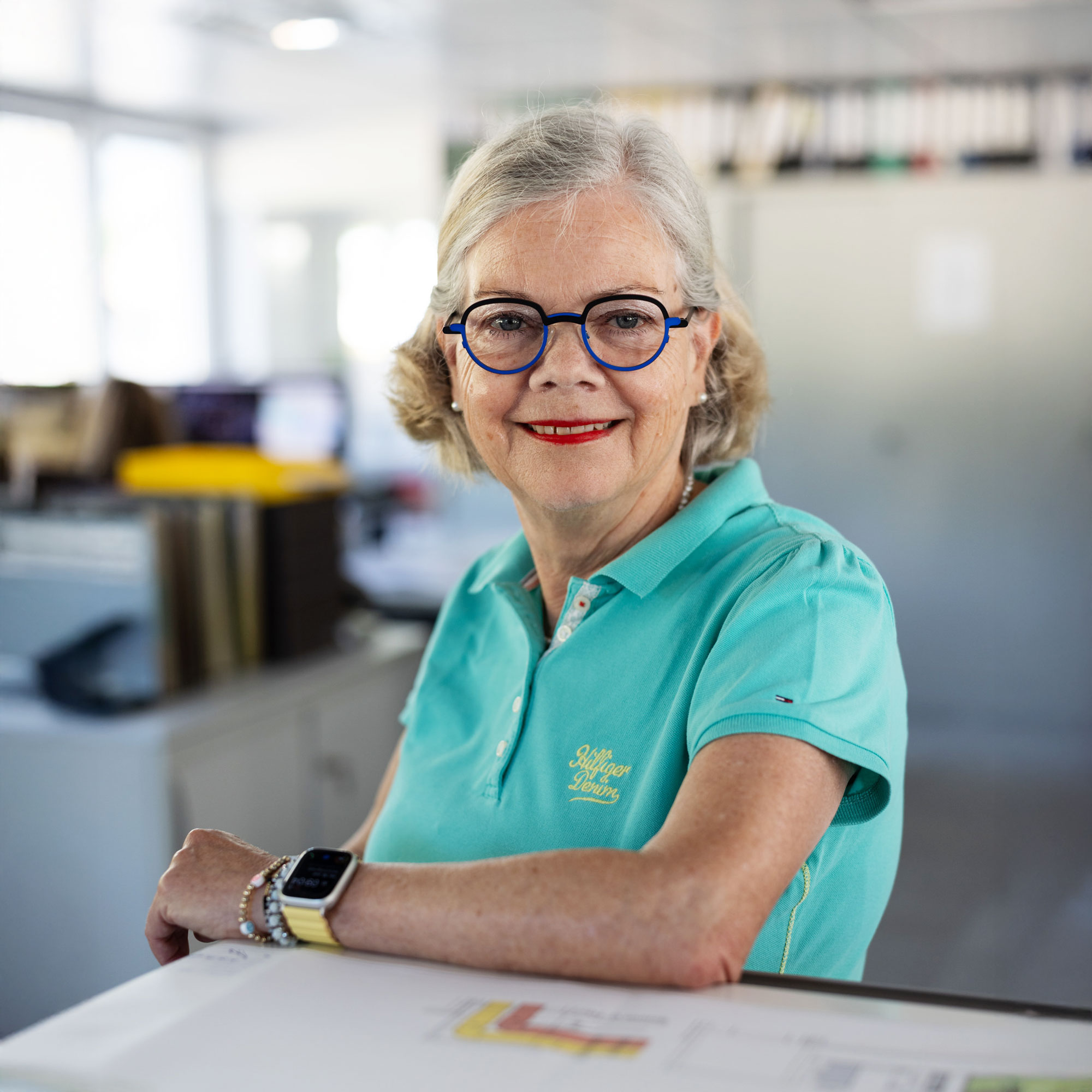 Frau im türkisfarbenen Shirt sitzt an einem Tisch und lächelt direkt in die Kamera